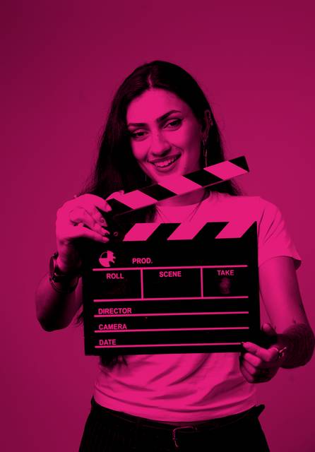 Una mujer sostiene una claqueta de cine con una sonrisa en un fondo rosa.