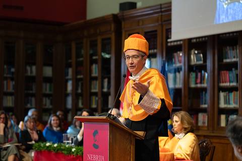 Un hombre con un traje académico naranja y un birrete habla desde un podio en un evento formal.