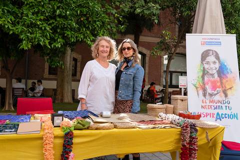 Dos mujeres posan juntas frente a una mesa decorada con artesanías y un cartel que invita a la acción social.