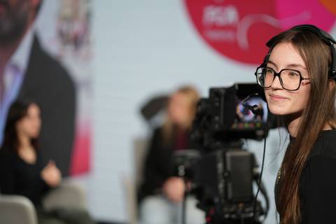 Una joven con gafas sonríe mientras opera una cámara en un escenario con un fondo desenfocado.