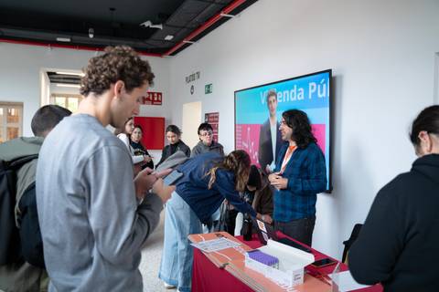 Un grupo de personas interactúa en un evento frente a una mesa con información y un panel detrás.