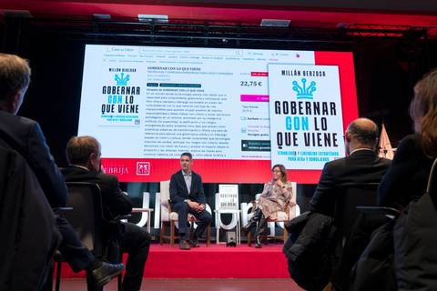 La imagen muestra una presentación de un libro en un escenario con un proyector y un público sentado.