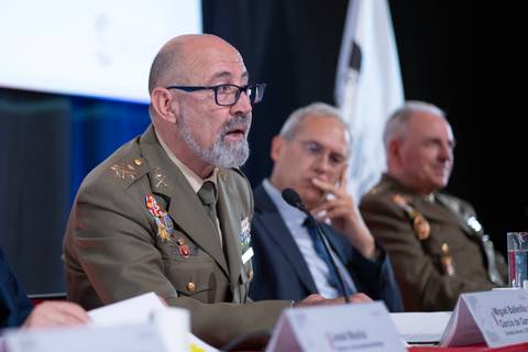 Un grupo de personas discute en una mesa de conferencias, con un oficial militar hablando al micrófono.