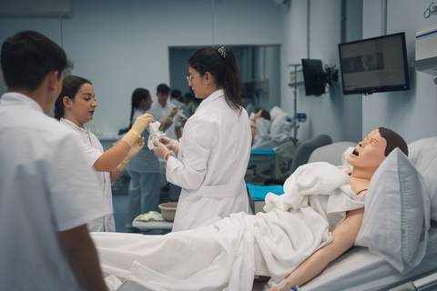 Un grupo de estudiantes en un laboratorio simulan un procedimiento médico con un maniquí en una cama de hospital.