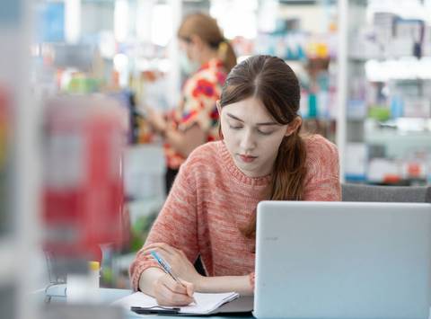 Una joven concentrada escribiendo en un cuaderno frente a una laptop en un entorno de farmacia.