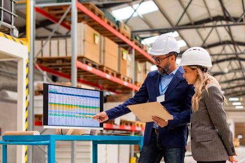 Un hombre y una mujer, ambos con cascos de seguridad, revisan datos en una pantalla de computadora en un almacén.