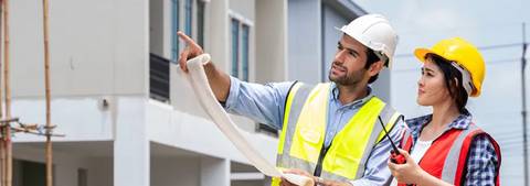 Un hombre y una mujer, ambos con cascos de seguridad, observan planos de construcción en un sitio de obra.