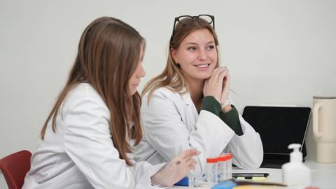 Dos mujeres jóvenes, vestidas de blanco, conversan y sonríen en un entorno de laboratorio.