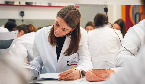 Una estudiante viste un bata blanca mientras estudia en un aula lleno de compañeros.