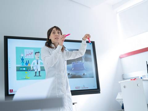 Una mujer en una bata de laboratorio presenta un experimento frente a una pantalla interactiva en un aula de ciencias.