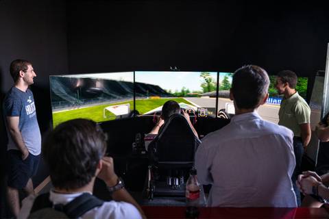 Un grupo de personas observa a un jugador en un simulador de carreras con pantallas múltiples.