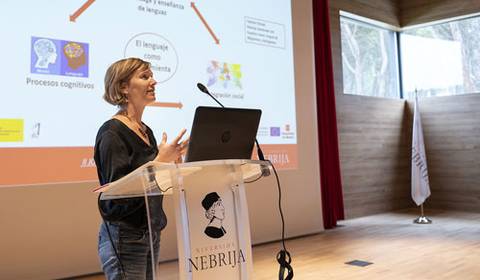 Una mujer presenta una charla en un auditorio, mientras interactúa con una pantalla que muestra conceptos relacionados con el lenguaje y los procesos cognitivos.