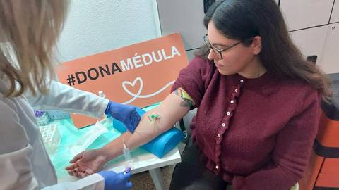 Una mujer se somete a una extracción de sangre mientras sostiene el brazo en una mesa con un cartel que promueve la donación de médula ósea.