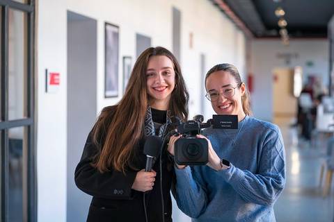 Dos jóvenes sonrientes posan en un pasillo con una cámara de video y un micrófono.