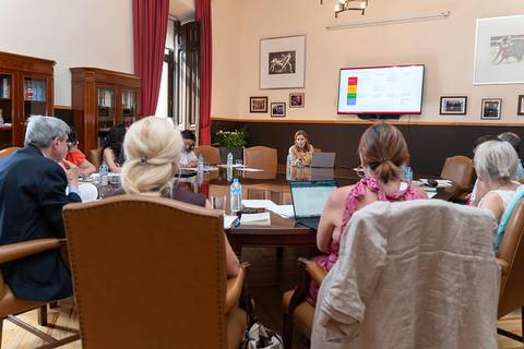Una reunión en una sala con varias personas sentadas alrededor de una mesa, mientras una mujer presenta información en una pantalla.