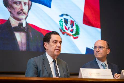 Dos hombres sentados frente a una pantalla que muestra un retrato antiguo y una bandera dominicana.
