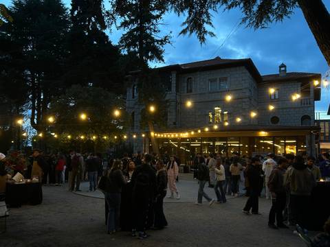 Una multitud de personas se reúne en un evento al aire libre, iluminado por luces colgantes, frente a un edificio en un ambiente nocturno.