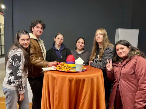 Un grupo de seis jóvenes posando juntos alrededor de una mesa con un mantel naranja y un sombrero de colores.