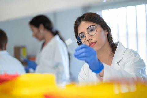 Una mujer en un laboratorio observa detenidamente un frasco mientras lleva guantes y gafas de seguridad.