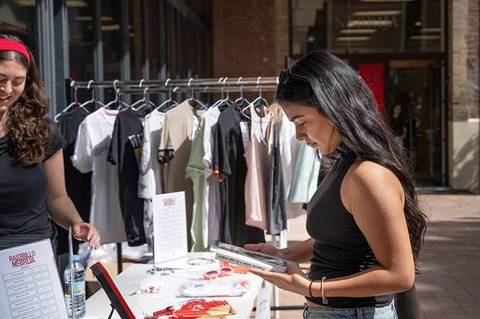 Dos mujeres están en un mercadillo, una revisando un producto mientras la otra le sonríe.