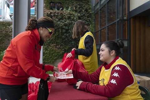 Varias personas están interactuando en un evento, donde una mujer entrega un ticket a otra en un stand con bolsas de color rojo.