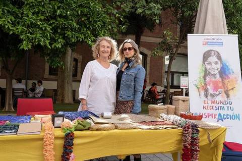 Dos mujeres posan juntas junto a una mesa decorada con artesanías y un cartel que invita a inspirar acción.