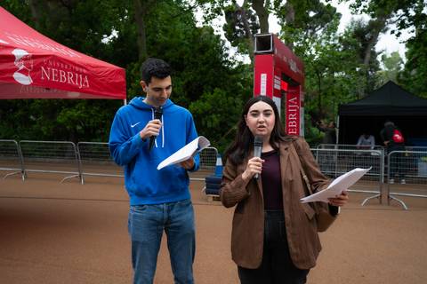 Dos presentadores están en un evento al aire libre, uno leyendo un papel y la otra hablando al micrófono.