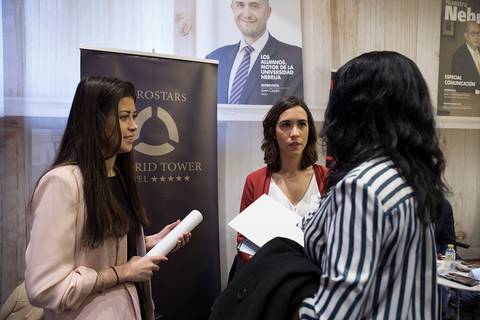 Tres mujeres están conversando en un evento o feria, rodeadas de carteles promocionales.