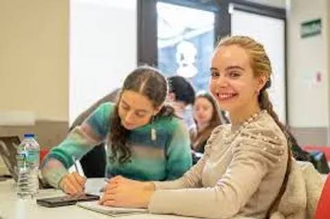 Dos jóvenes sentadas en una mesa de estudio, una sonríe mientras la otra escribe en un cuaderno.