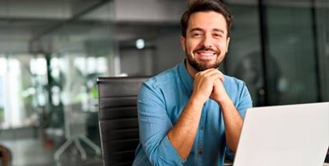 Un hombre sonriente sentado frente a una computadora portátil en un entorno de oficina moderno.