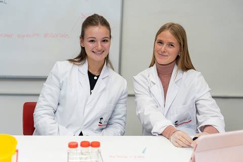 Dos jóvenes sonrientes con batas de laboratorio posan en un entorno académico.