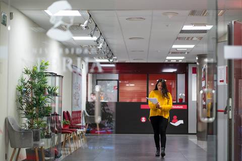 Una mujer camina por un pasillo luminoso de oficina mientras sostiene unos documentos.