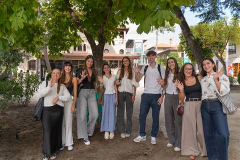 Un grupo de diez jóvenes posando con sonrisas y gestos amistosos en un parque, rodeados de árboles y edificios coloridos.