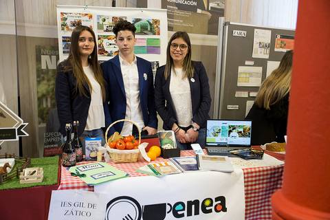 Tres jóvenes participan en una feria escolar mostrando su proyecto en un stand decorado.