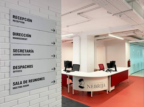 Una vista interior del Instituto Nebrija con señalización de diferentes áreas y una recepción.