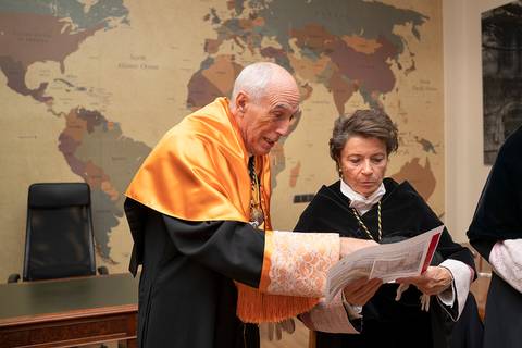Dos personas con vestimenta académica revisan un documento frente a un mapa mundial.