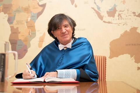 Un hombre con toga azul y pajarita está escribiendo en un libro frente a un mapa del mundo.