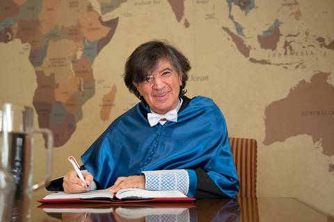 Un hombre con una capa azul y un moño escribe en un libro frente a un mapa del mundo.