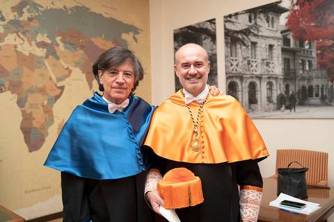 Dos hombres en trajes académicos posan juntos en un ambiente interior con un mapa del mundo en la pared.