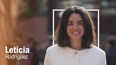 Una joven sonriente con cabello oscuro, vestida con un abrigo claro, se encuentra en un entorno urbano.