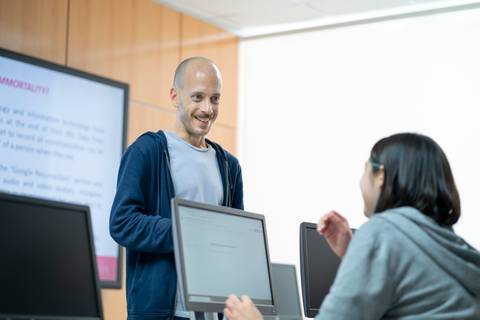 Un hombre sonriente interactúa con una mujer en un salón de clases, rodeados de computadoras.