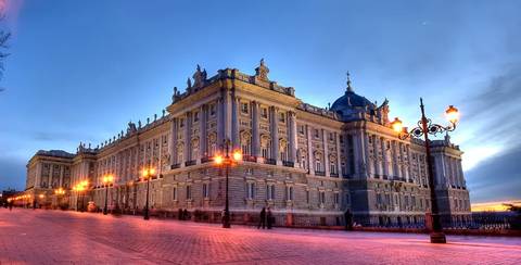 El Palacio Real de Madrid se muestra iluminado al atardecer con un cielo azul de fondo.