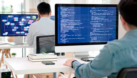 La imagen muestra a dos personas trabajando en un entorno de oficina con pantallas de computadora que muestran código y gráficos