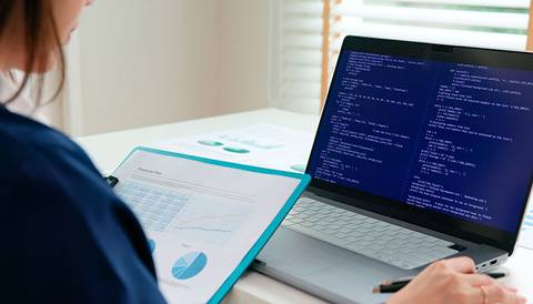 La imagen muestra a una persona revisando gráficos en una hoja mientras trabaja en una computadora portátil con código en la pantalla.