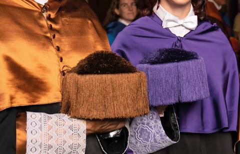 Dos personas con capas y birretes de diferentes colores en un evento ceremonioso.
