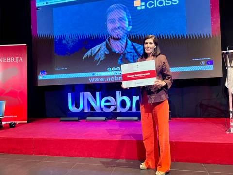 Una mujer sonriente sostiene un cartel mientras aparece un video de otra persona proyectado de fondo en un evento.