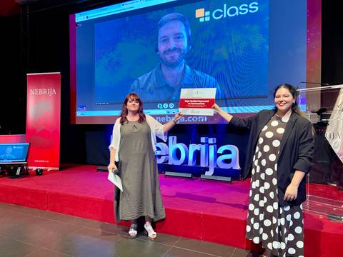 Dos mujeres presentan un reconocimiento en un evento con un fondo digital donde aparece un hombre en video.