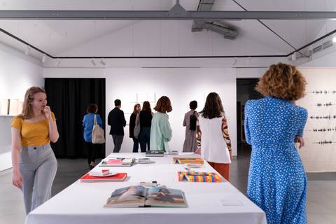 Una inauguración en una galería de arte donde varias personas observan las obras mientras se encuentran en una mesa con libros y materiales de exposiciones.