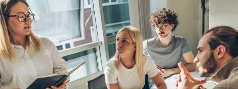 Un grupo de personas en una reunión escuchando atentamente a una mujer que presenta información con un cuaderno en la mano.