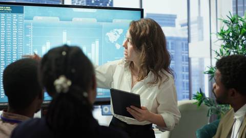 Una mujer presenta información en una pantalla a un grupo de personas en un entorno de oficina moderna.